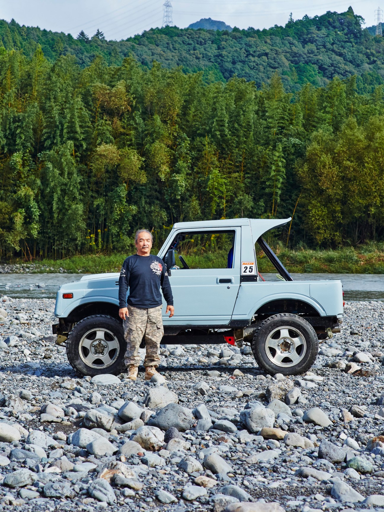 Yasumasa Ishikawa standing next to his modified white Suzuki Jimny on rocky riverbank with forested hills