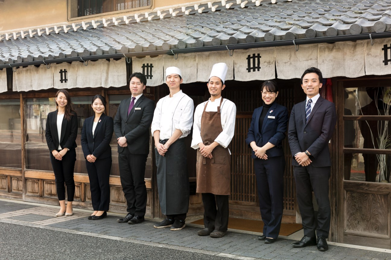 Seven staff members in formal attire standing outside a traditional Japanese building with tiled roof in Sasayama.