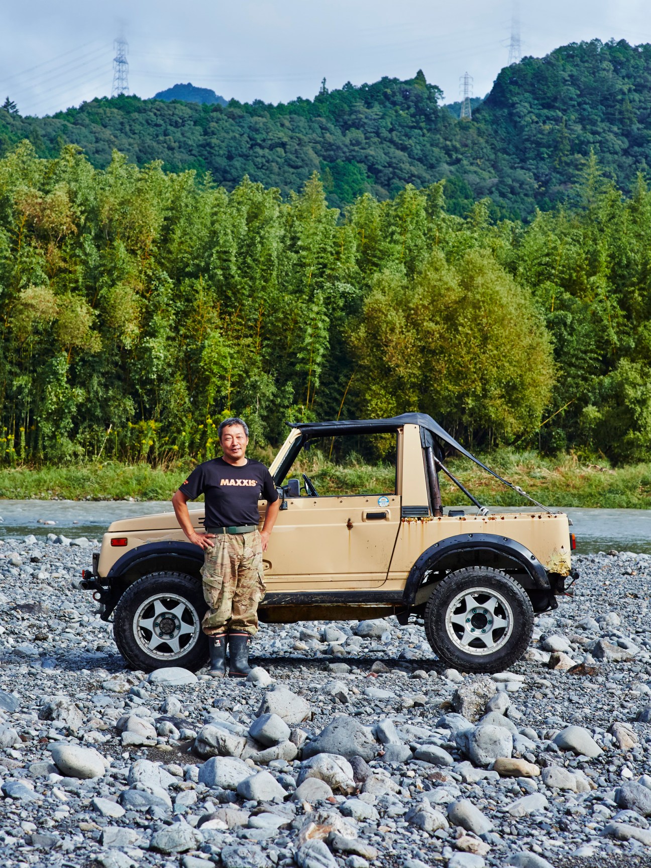 Koichi Yoshida standing beside his beige SJ40 Suzuki Jimny on a rocky riverbank with forested hills in background