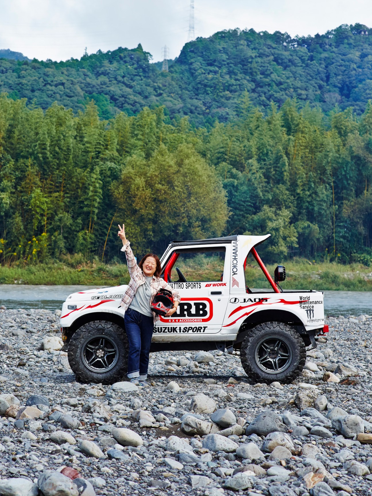 Noriko Ito making peace sign next to white Suzuki Jimny on rocky riverbank with forested hills in background