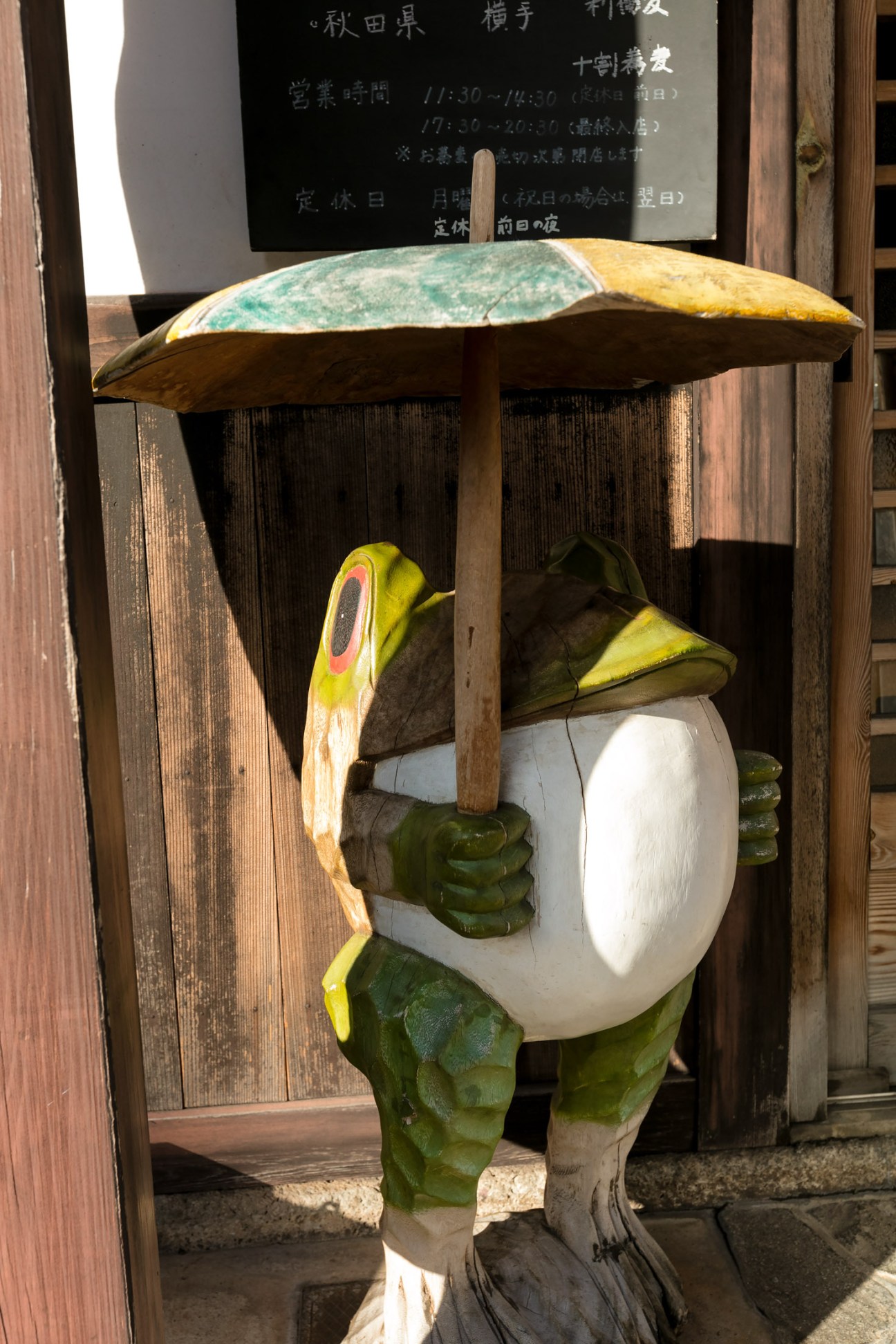 Wooden statue of a smiling figure in green and yellow holding an umbrella outside a Japanese shop.