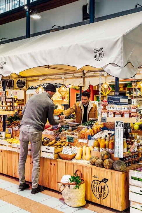 A vendor serves a customer at a market stall selling fresh produce and local goods under a white awning.