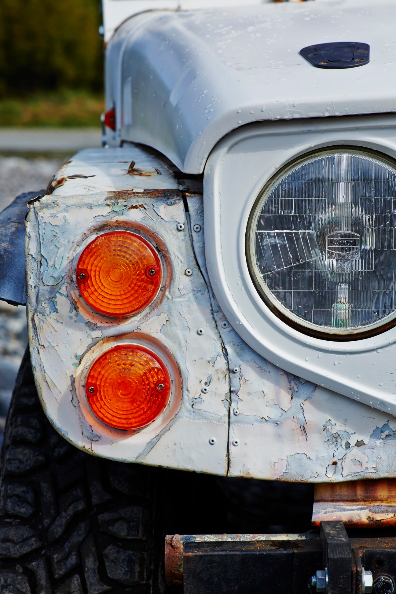 Close-up of weathered white Suzuki Jimny showing round headlight and orange turn signals with peeling paint