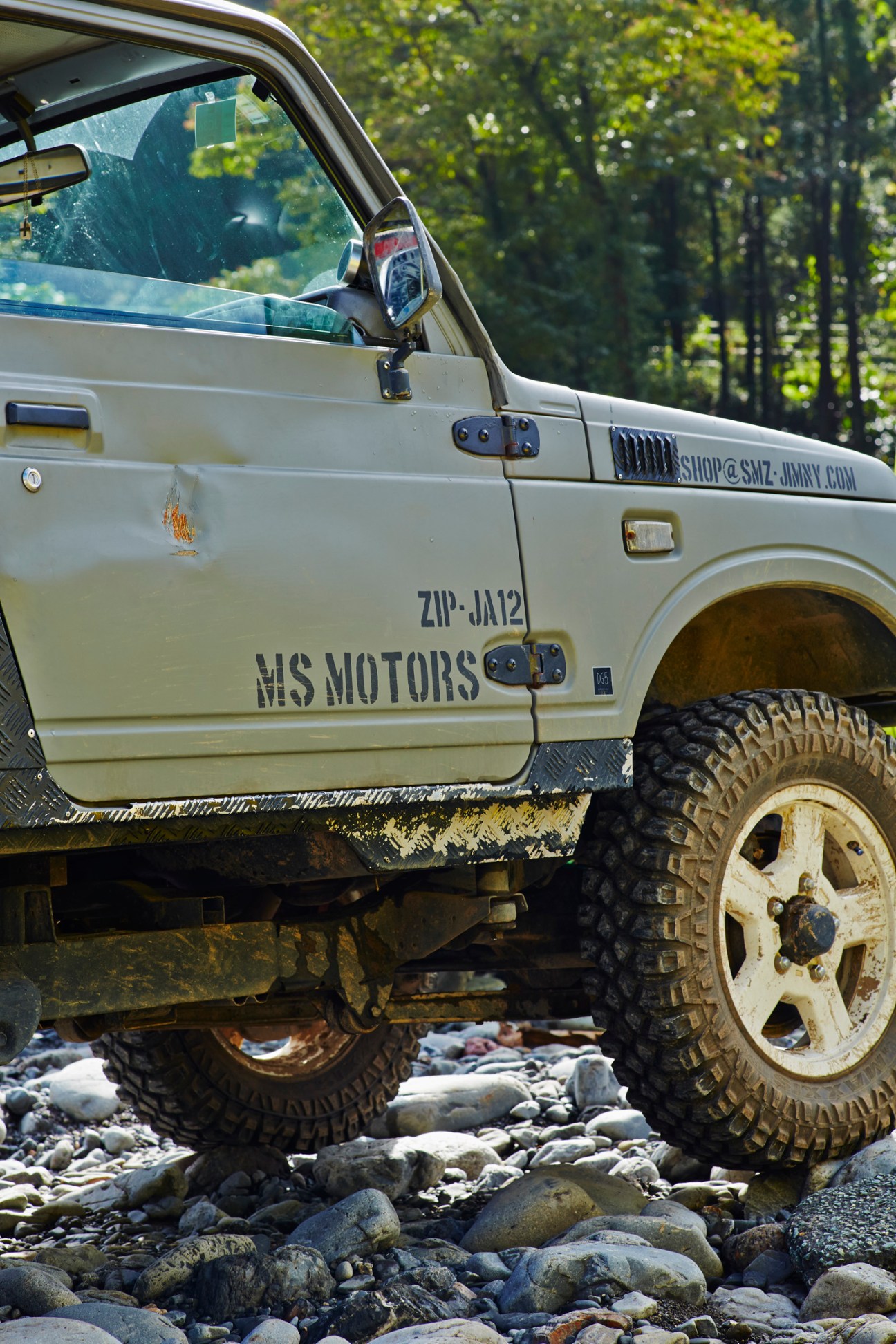 Modified Suzuki Jimny with MS Motors branding navigating rocky riverbank terrain at off-road event