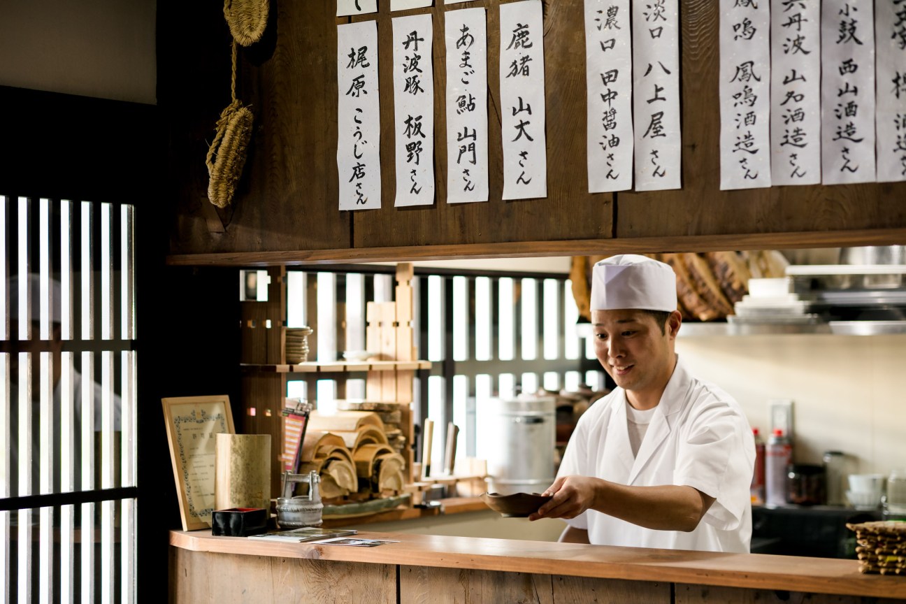 Chef Tomoaki Maekawa in traditional Japanese restaurant kitchen with white chef's uniform and hat.