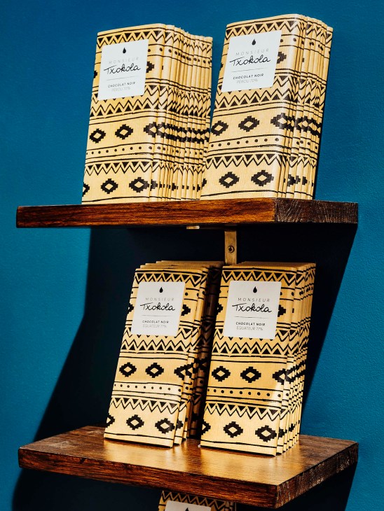 Monsieur Txokola chocolate bars with geometric patterned packaging displayed on wooden shelves against a blue background.