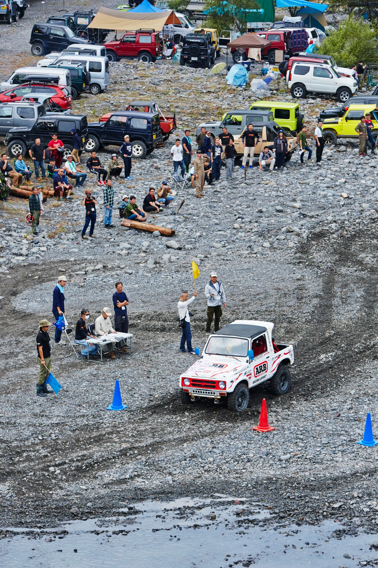 White Suzuki Jimny with racing number navigating rocky terrain at competitive event with spectators and vehicles