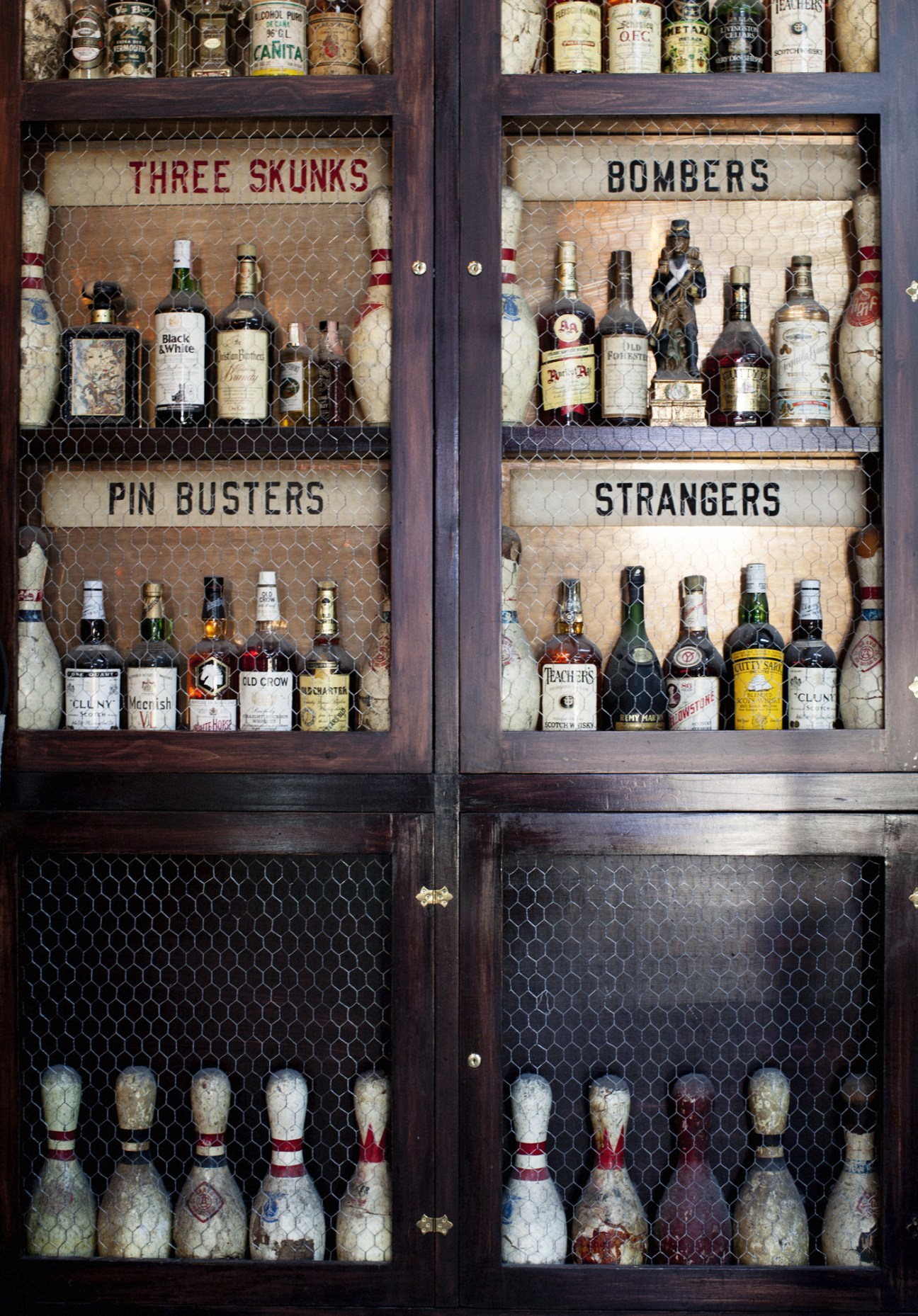 Vintage wooden cabinet display with glass doors containing labeled sections of spirits and cocktail ingredients.