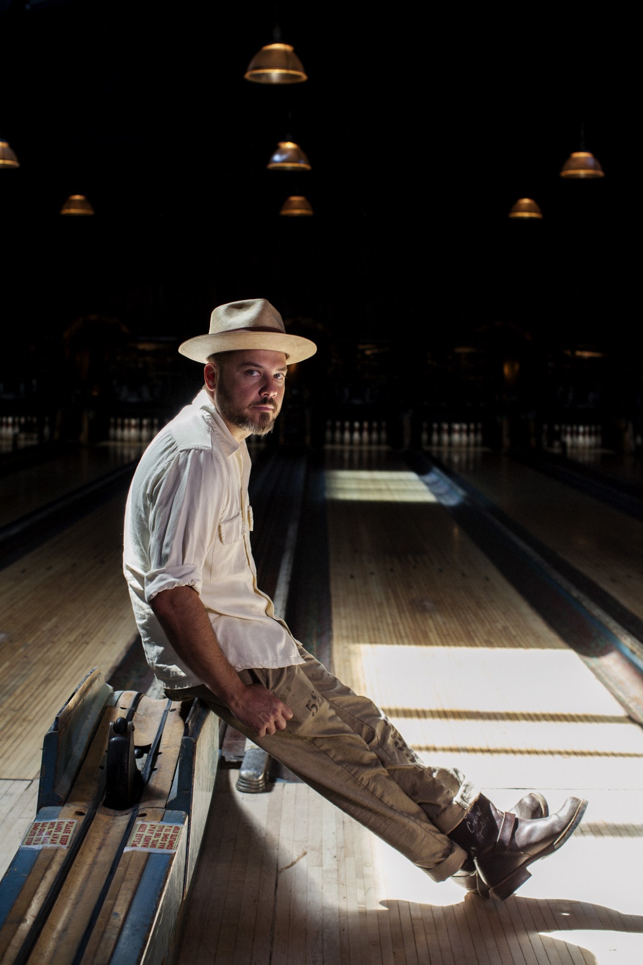 Bobby Green poses on a bowling lane at Highland Park Bowl, wearing a white shirt and tan hat under brass pendant lights.