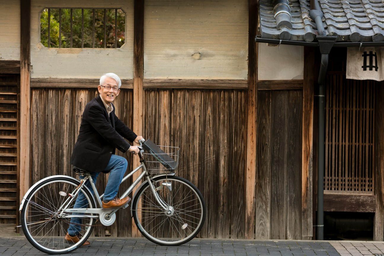 Community leader Toshiaki Imamura smiles while sitting on a bicycle in front of a traditional wooden building in Sasayama, Japan.