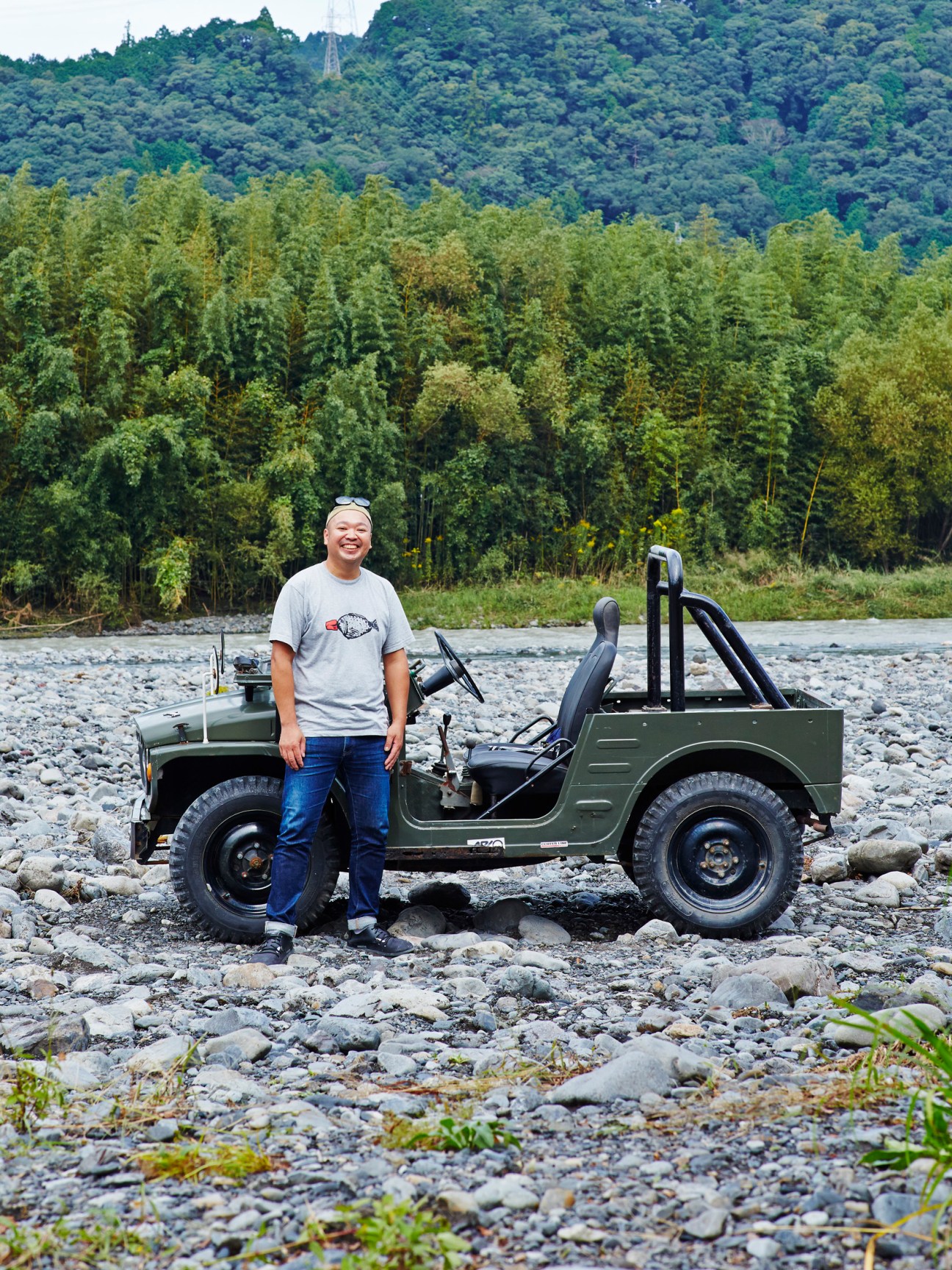 Hiroki Nakamura standing beside his vintage olive green LJ20 Suzuki Jimny on rocky riverbank