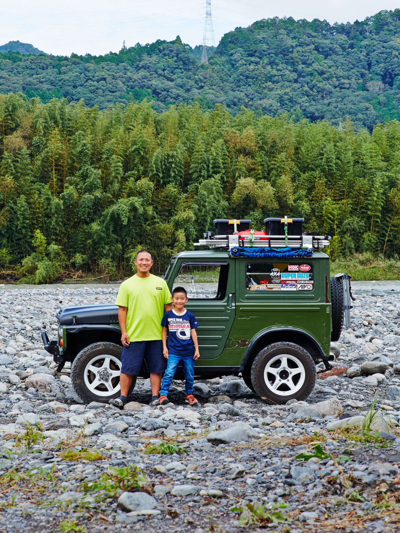 Father and son Toshihiko Muramatsu and Yuha standing beside their green DIY sleeper Suzuki Jimny on rocky riverbank