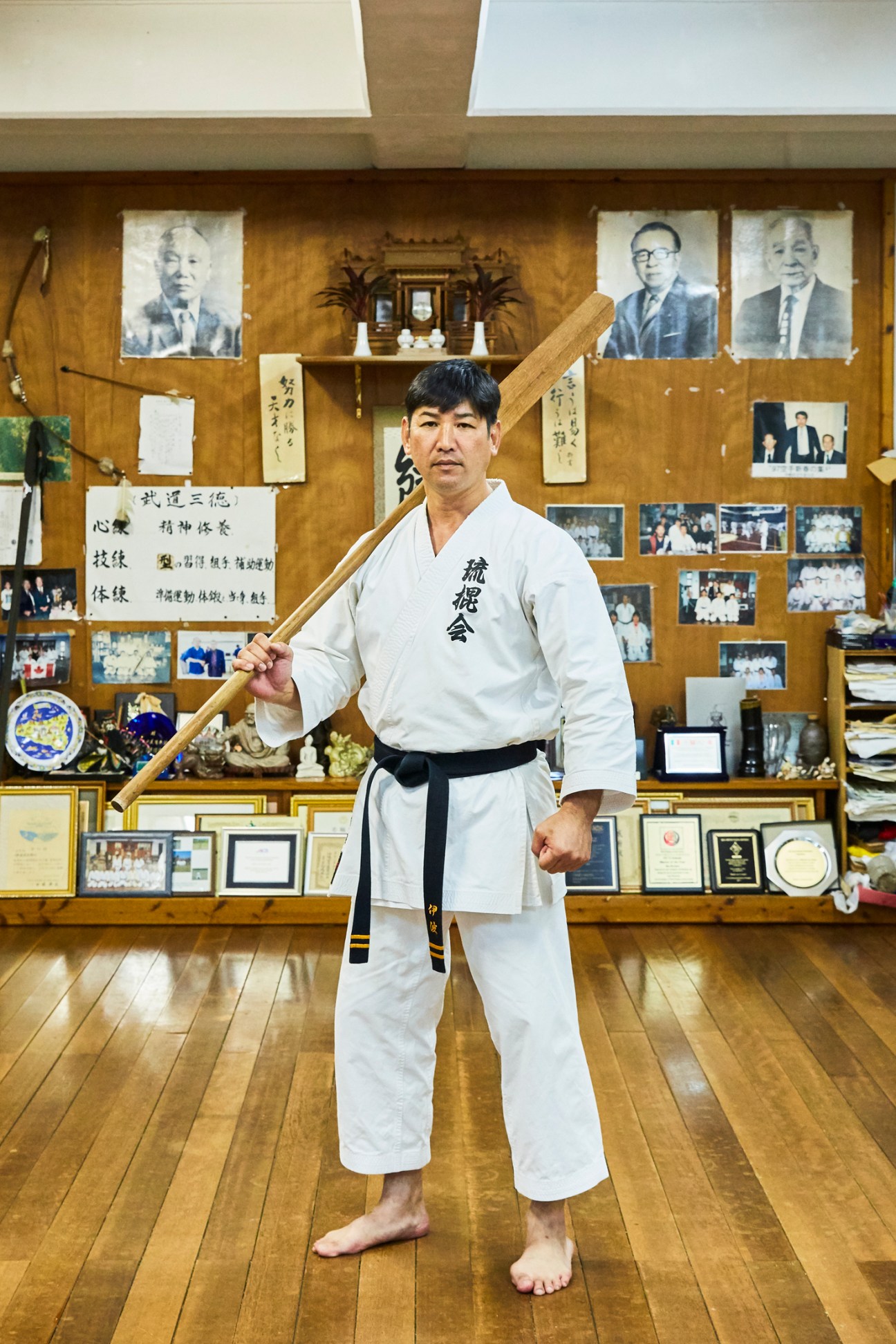 Mitsutada Iha in white martial arts uniform holding wooden staff in traditional dojo with portraits and awards