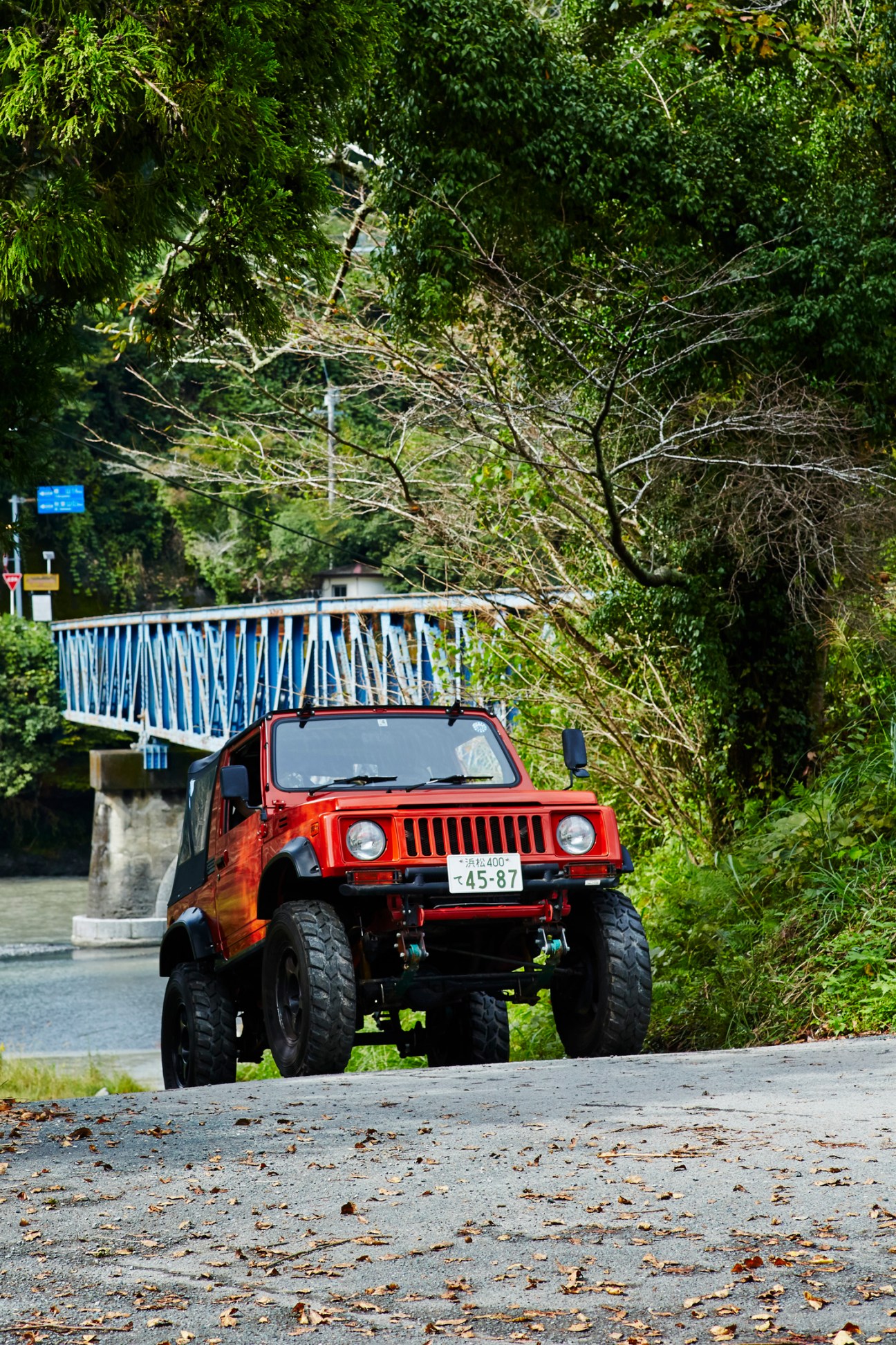 Red Suzuki Jimny SUV parked on rural Japanese road with bridge and lush green vegetation in background