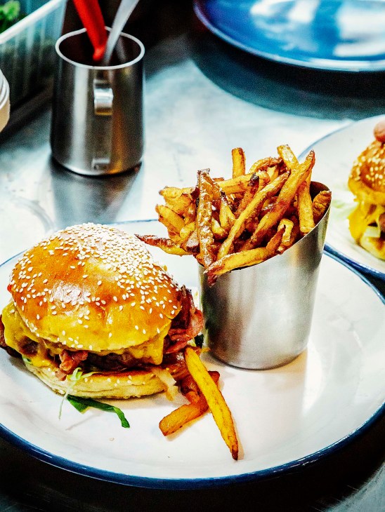 A burger with sesame seed bun and bacon topped with melted cheese, served with golden fries in a metal cup on a white plate with blue rim.