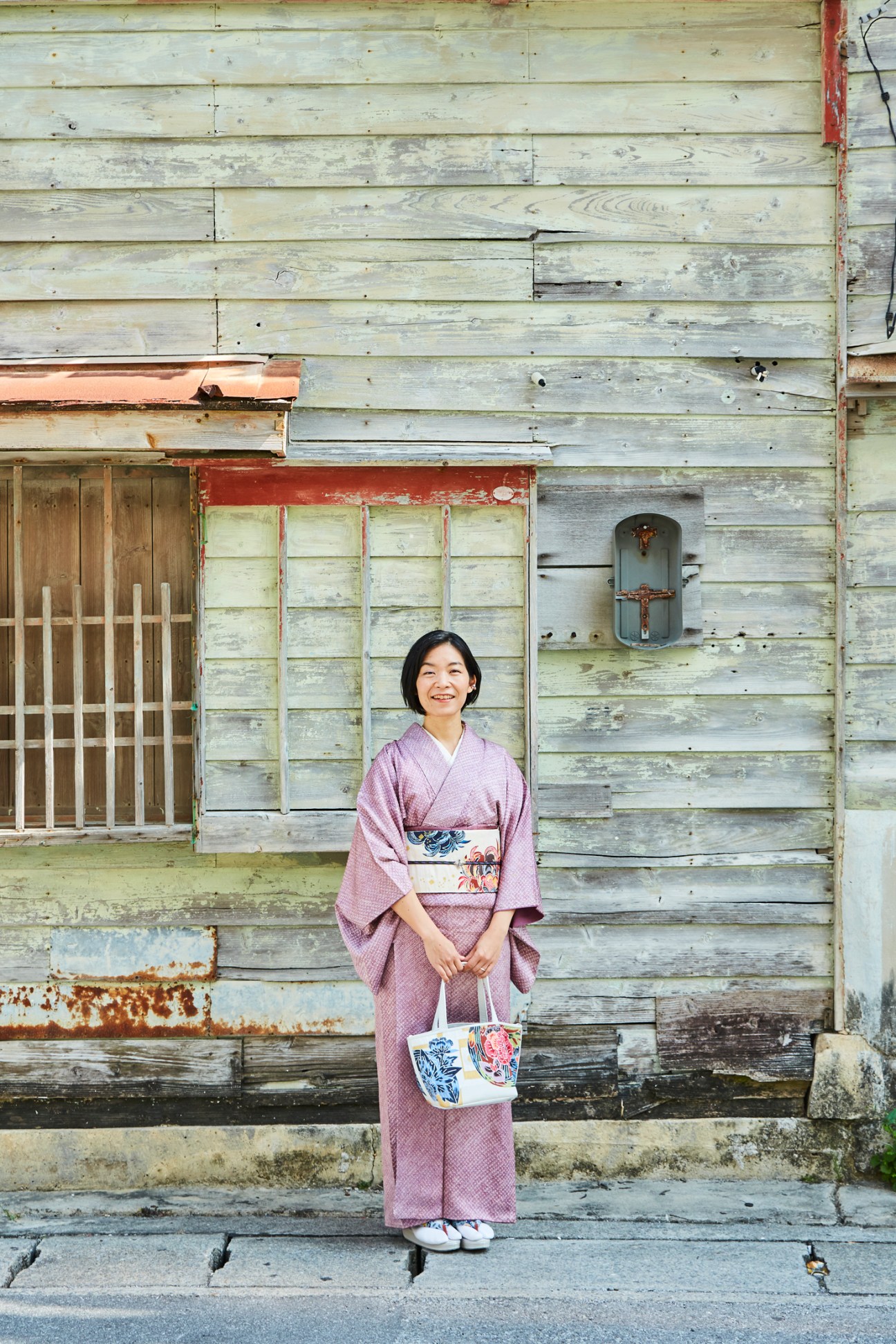 Tomoko Nawa, an Okinawan bingata dye artist, wearing a purple kimono and standing in front of weathered wooden building