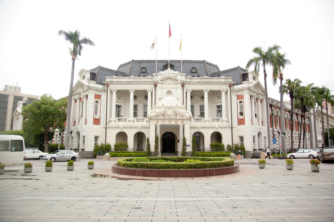 White neoclassical government building with columns, slate roof, and flags, fronted by a landscaped plaza with palm trees.