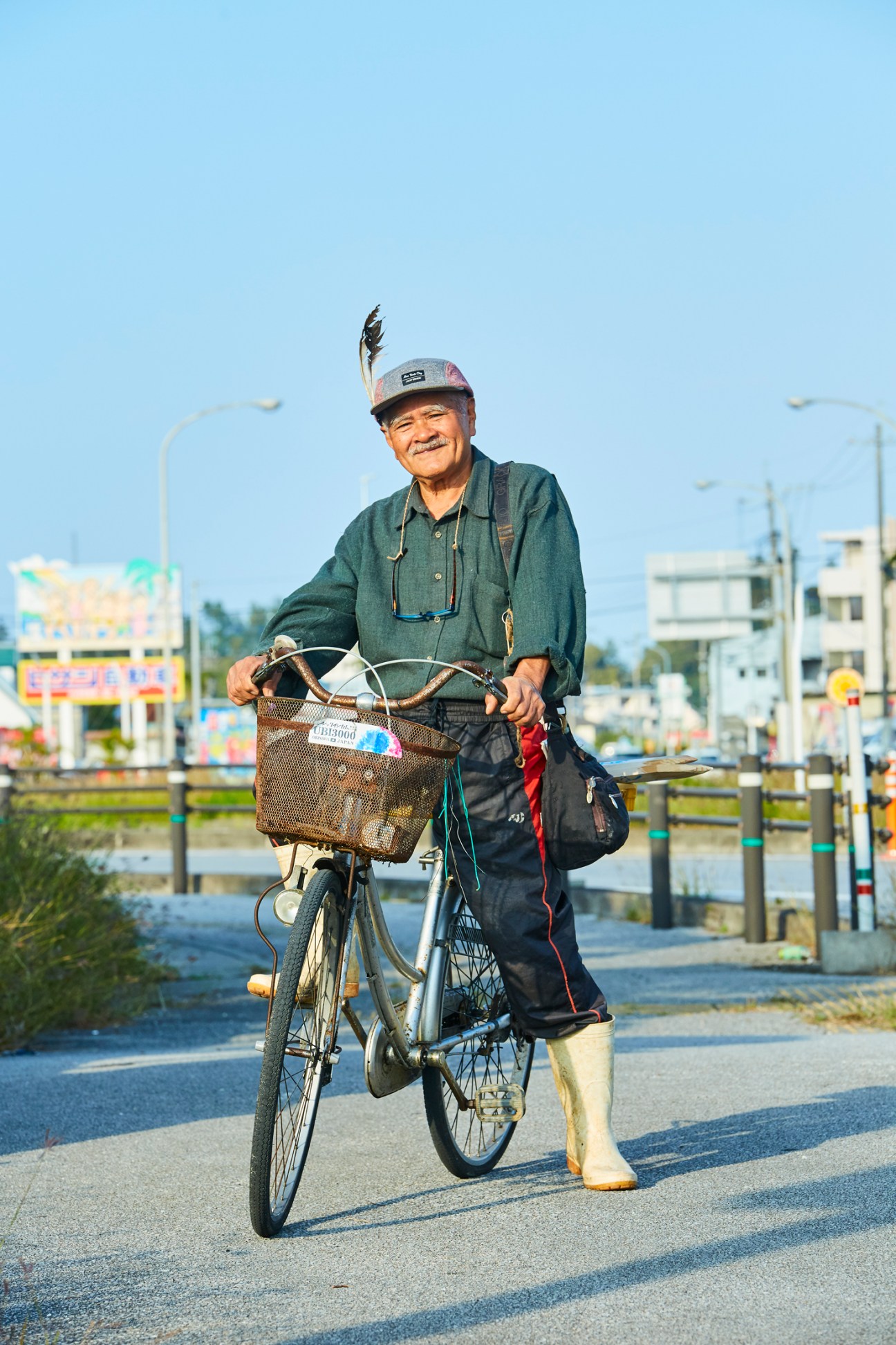 Elderly man with feathered cap standing beside bicycle on road in Okinawa