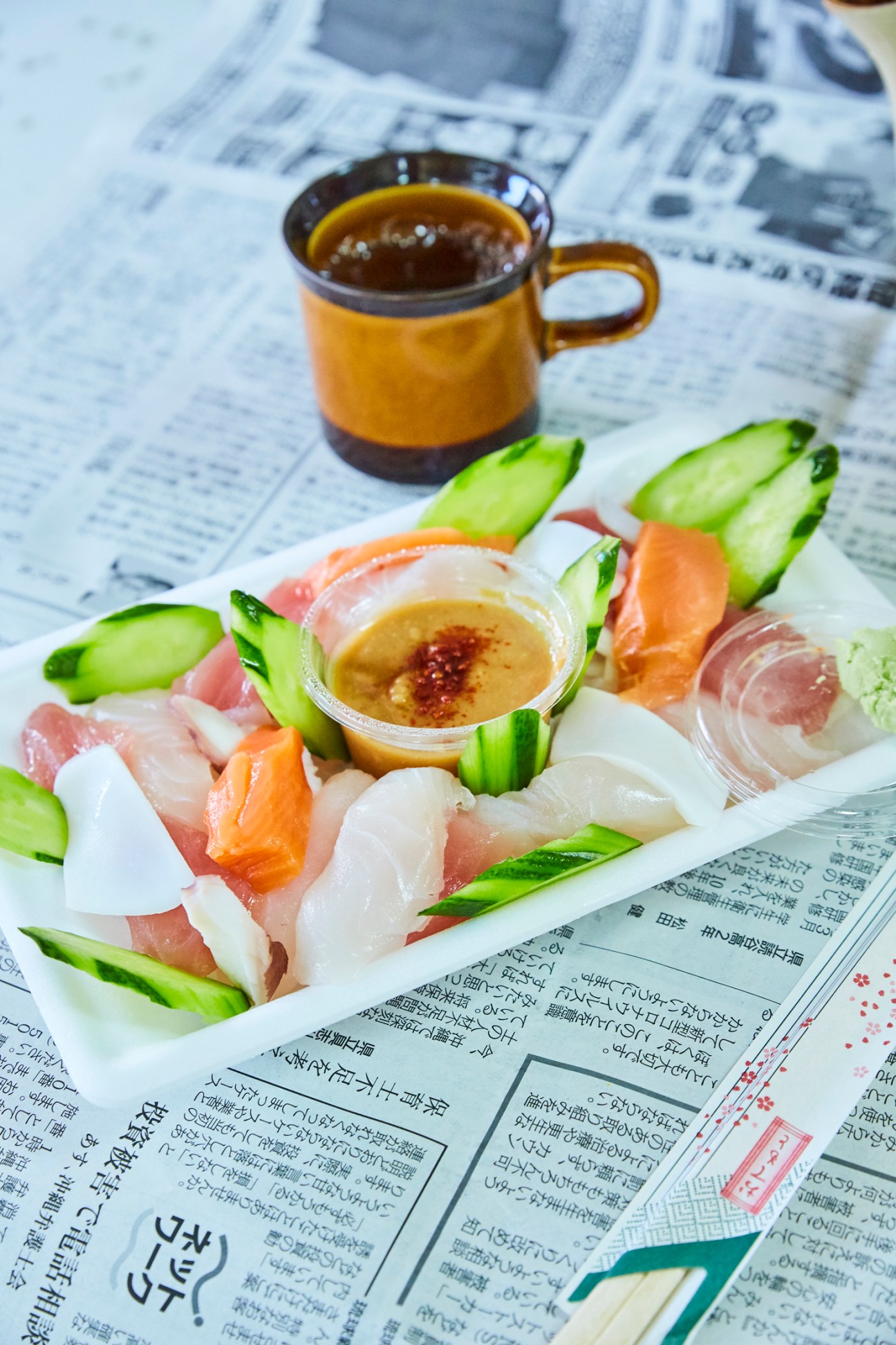 Fresh sashimi platter with sliced raw fish, cucumber garnish and dipping sauce served on newspaper