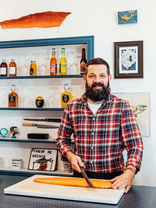 Man in red plaid shirt at his smoked-fish épicerie, holding a smoked fish on a cutting board with shelves of bottles behind him.