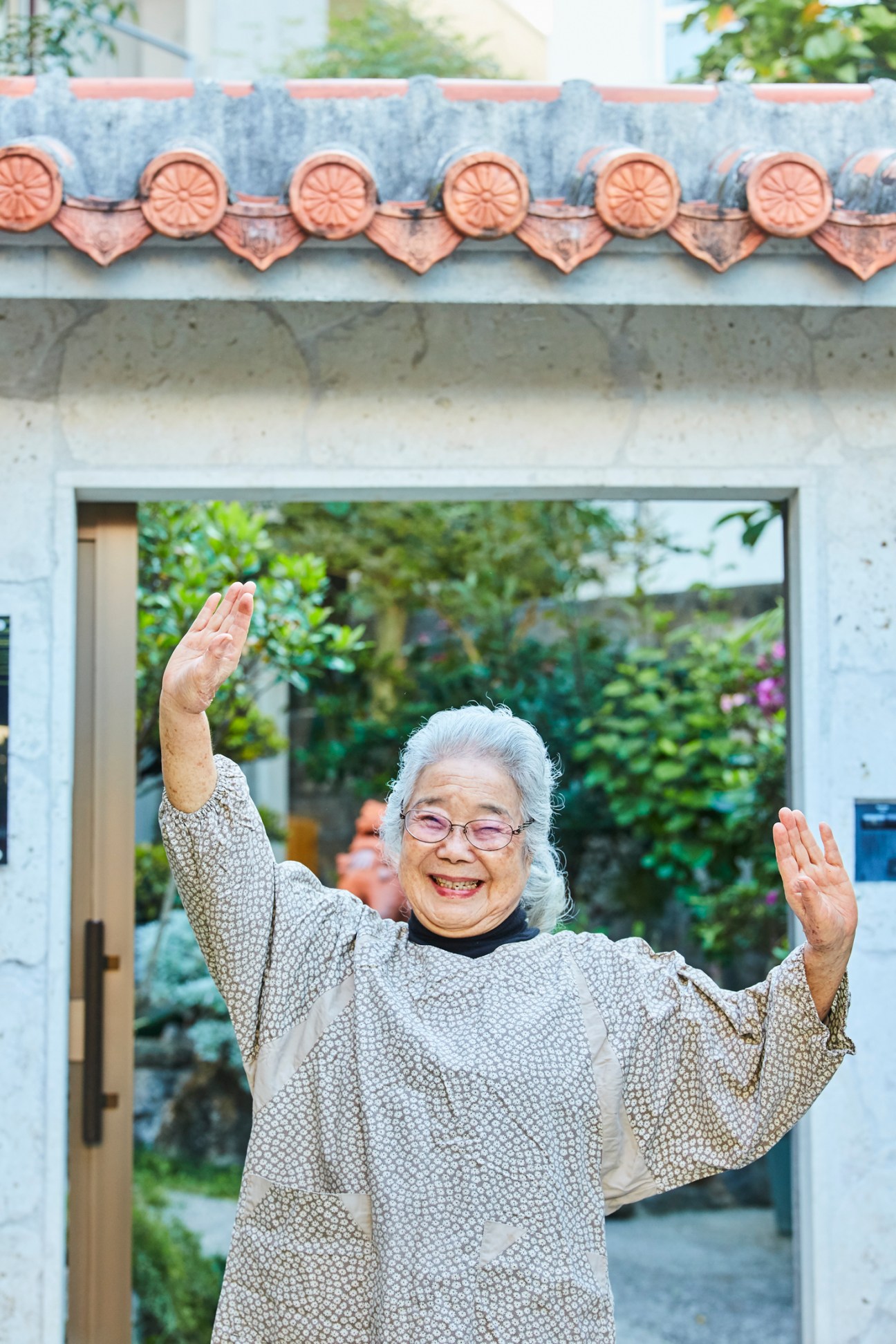 Elderly woman in patterned sweater and glasses raising both hands with joyful expression outside traditional building