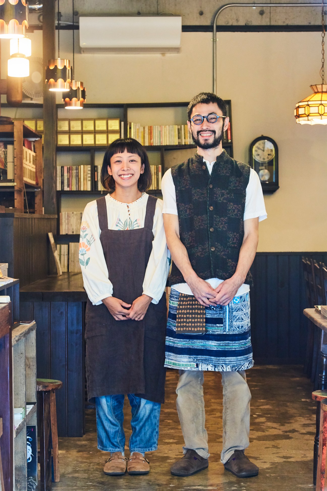 Akio and Aoi Yamazaki standing together at Mahou Coffee in Naha, wearing aprons in a cozy bookshelf-lined interior