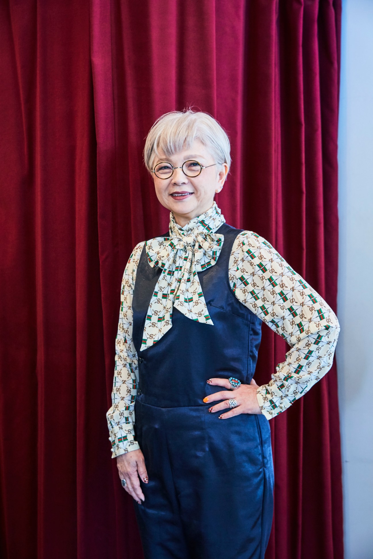 Woman with short blonde hair and round glasses wearing patterned blouse and navy vest posing with hand on hip