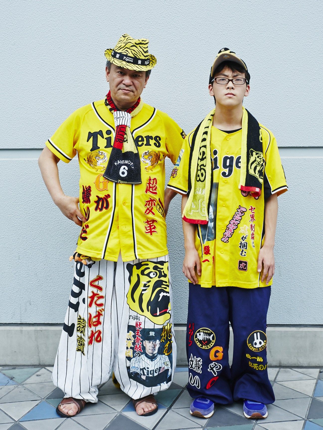 Two Hanshin Tigers fans in yellow uniforms decorated with Japanese characters and tiger logos stand against a white wall.