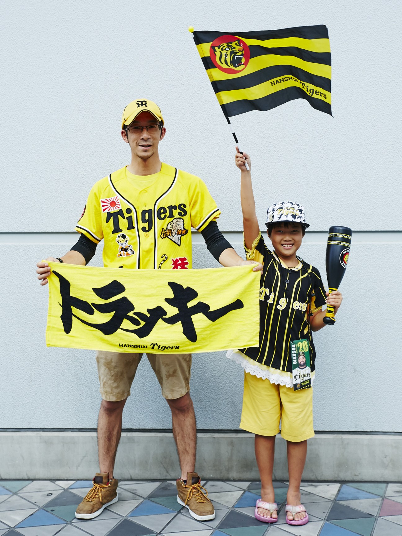 An adult and child in Hanshin Tigers gear holding flags and banners at a baseball stadium.