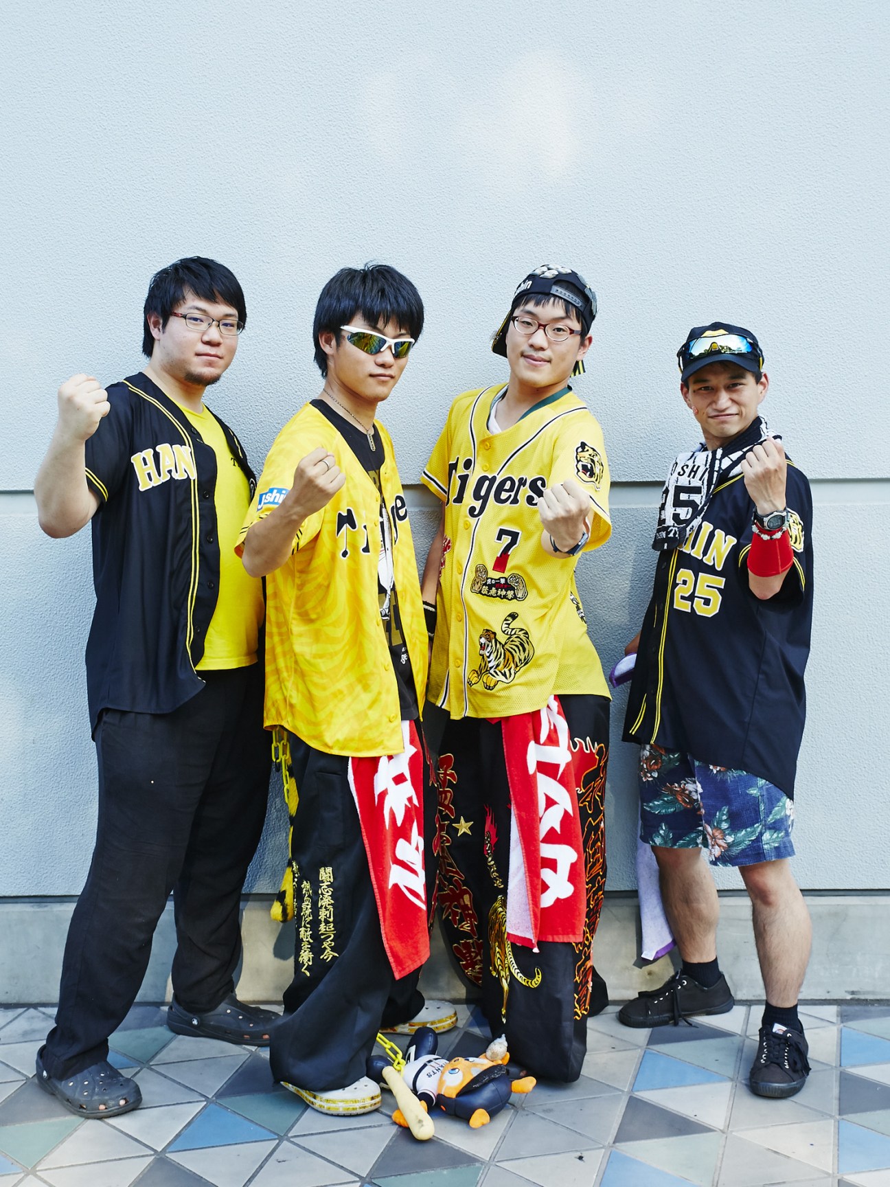 Four young male Hanshin Tigers fans posing together in yellow and black team uniforms and caps against a light blue wall.