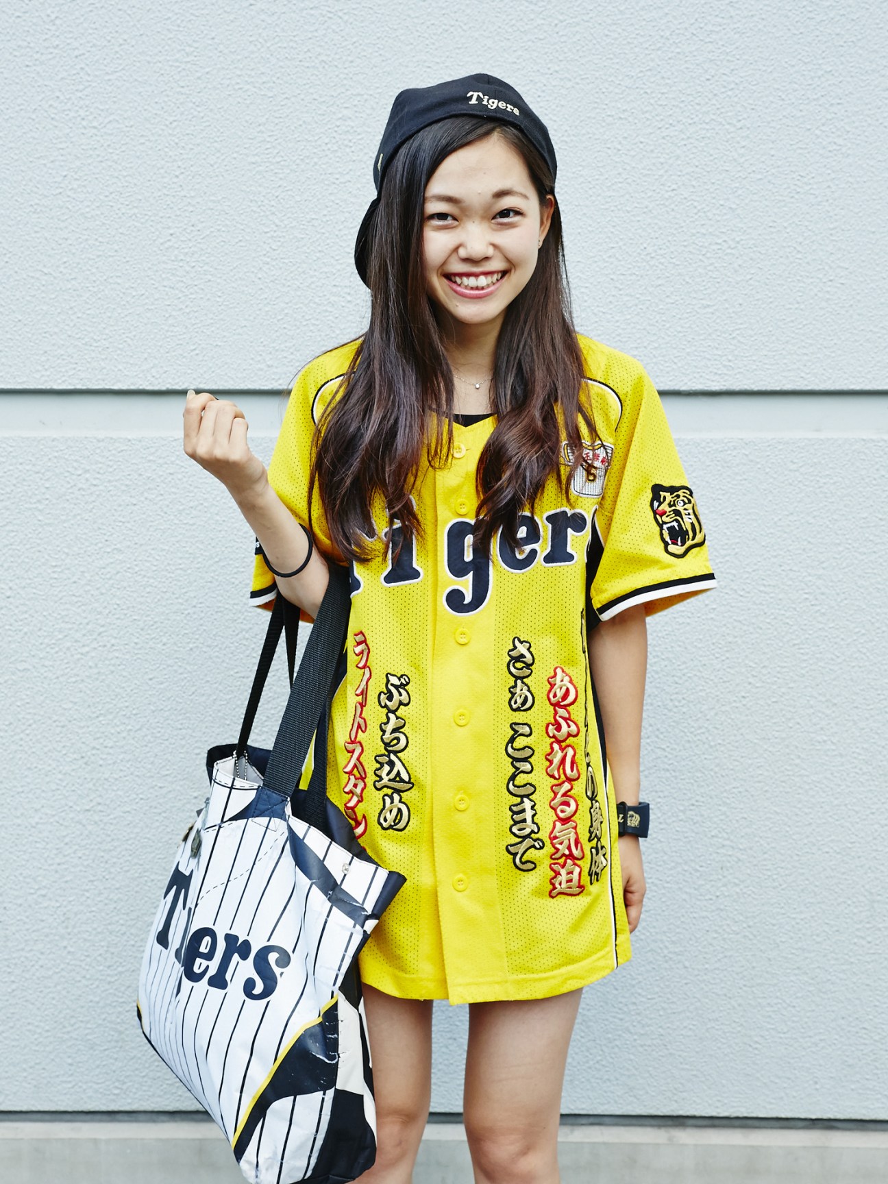 Young woman wearing a yellow Hanshin Tigers baseball uniform with Japanese characters and a Tigers cap, holding a pinstriped team bag.