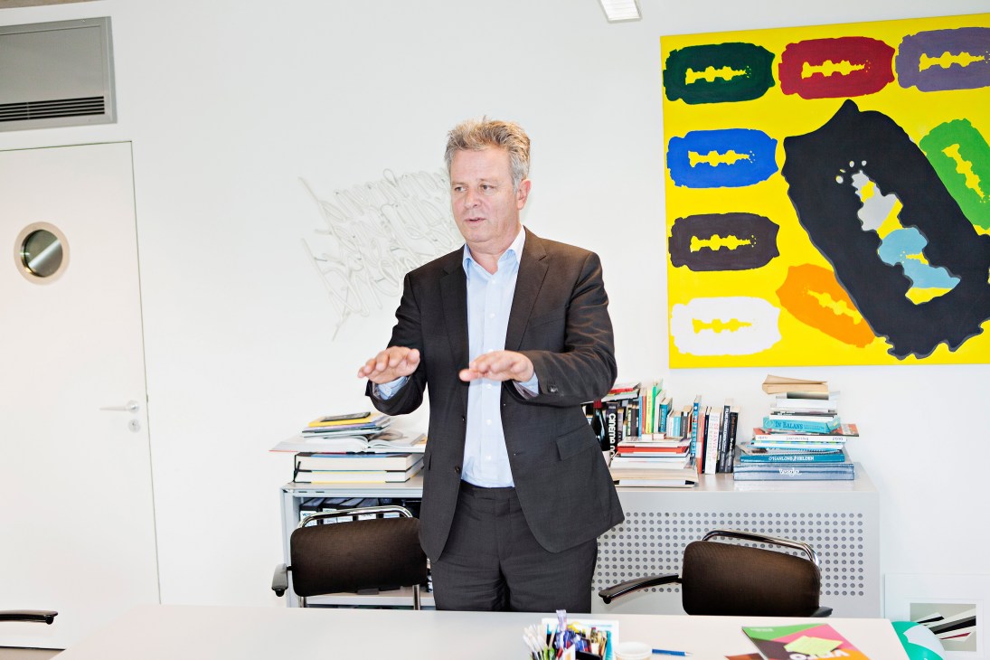 Managing director Lennart van der Meulen stands in his office gesturing while speaking, with stacks of books and colorful artwork visible behind him.