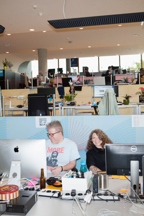 Two people work at computers in a modern office with a colorful sunburst mural behind them.