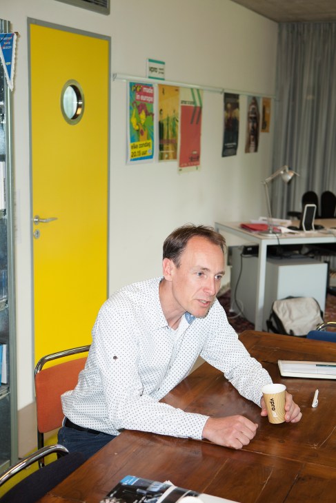 Stan van Engelen, VPRO's editor in chief of the TV department, sits at a wooden table holding a coffee cup in an office with a bright yellow door.
