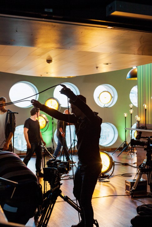 Film crew recording musicians with hurdy-gurdy and clarinet in studio with illuminated circular windows.