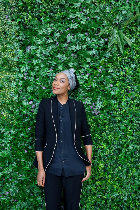 Singer Giovanca Ostiana stands against a lush green living wall with purple flowers.