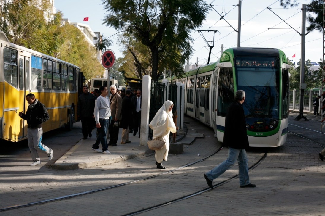 Modern green and white tram train from Alstom stops at a street crossing in Tunis with passengers and a yellow bus nearby.