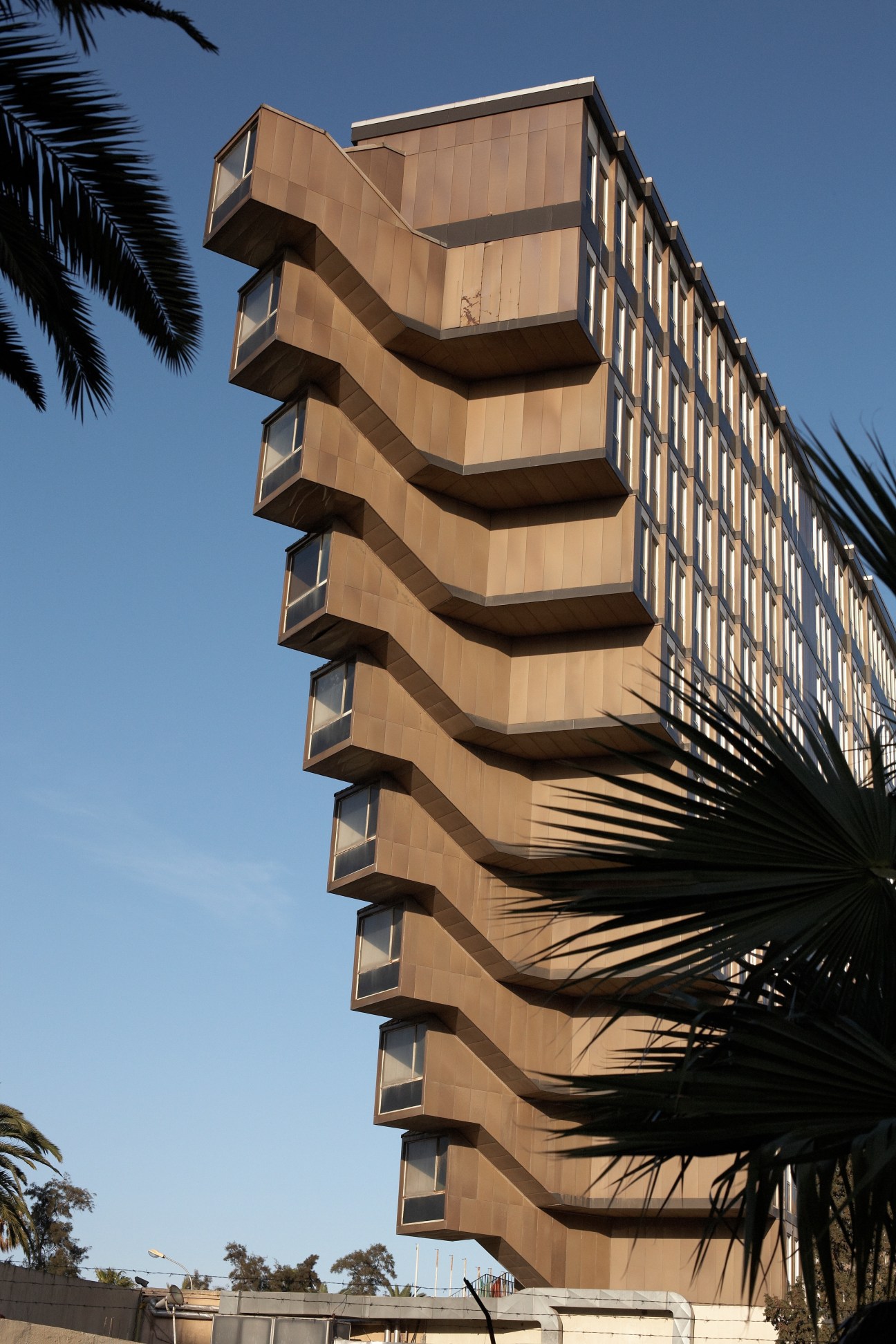 Hotel du Lac, a multi-story tan building with distinctive stacked balconies against a blue sky and palm fronds.