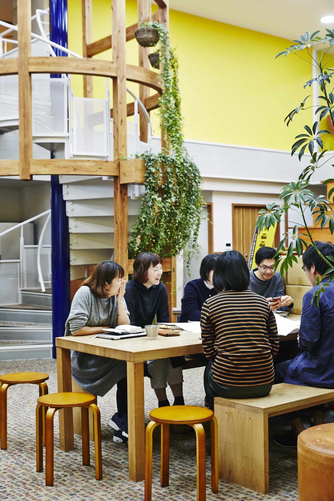 Six Hobonichi staff members gather around a wooden table in a bright yellow meeting room with climbing plants and wooden house-shaped booths.