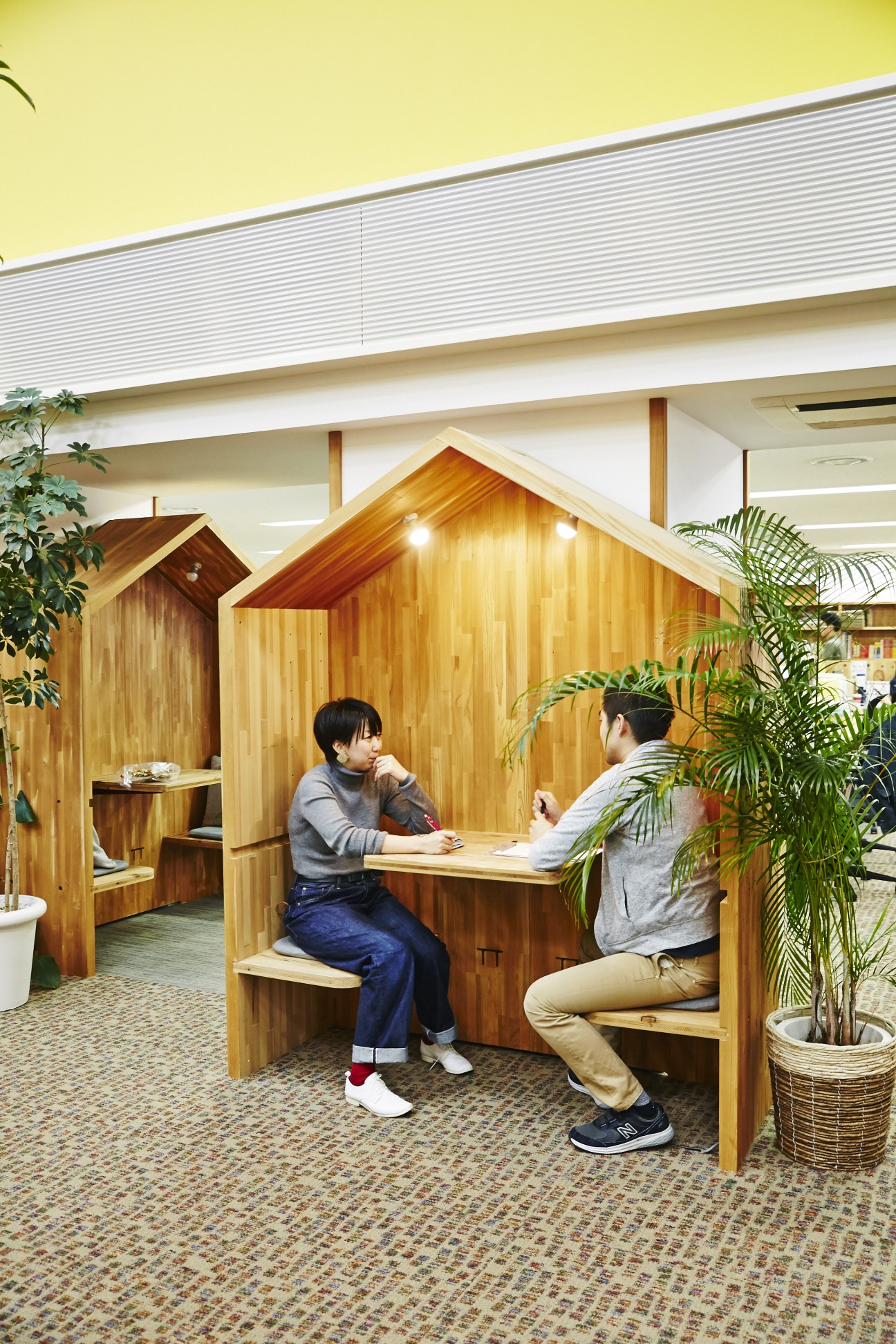 Two people sit inside wooden house-shaped booths at Hobonichi headquarters with plants nearby.