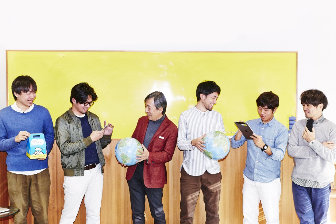 Shigesato Itoi demonstrates the Hobonichi Earthball to staff members against a yellow wall.