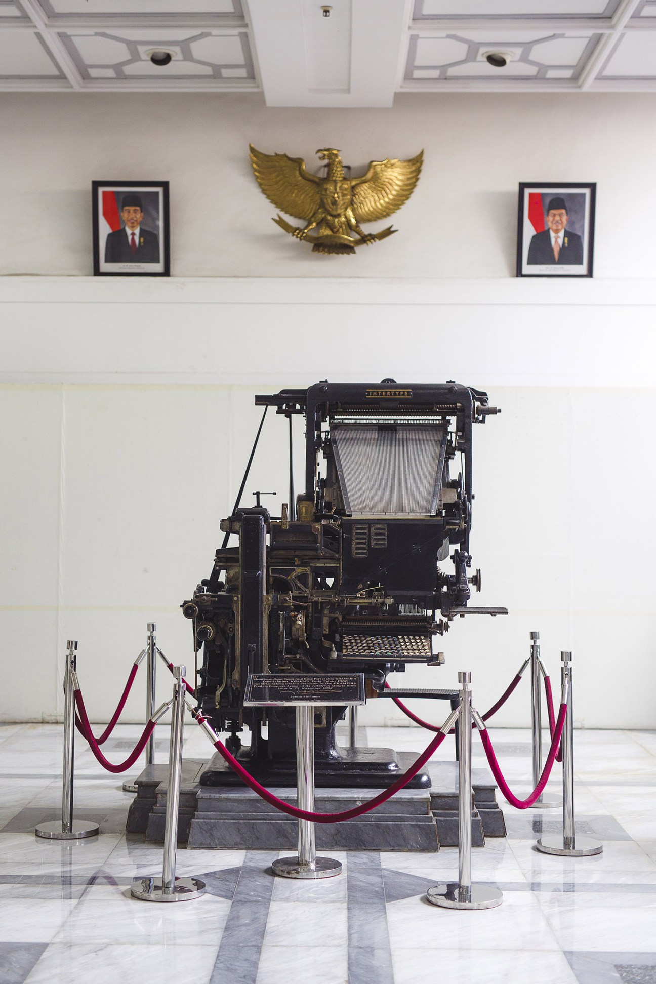 Vintage printing press display in Kompas Gramedia lobby with Indonesian eagle emblem and presidential portraits.