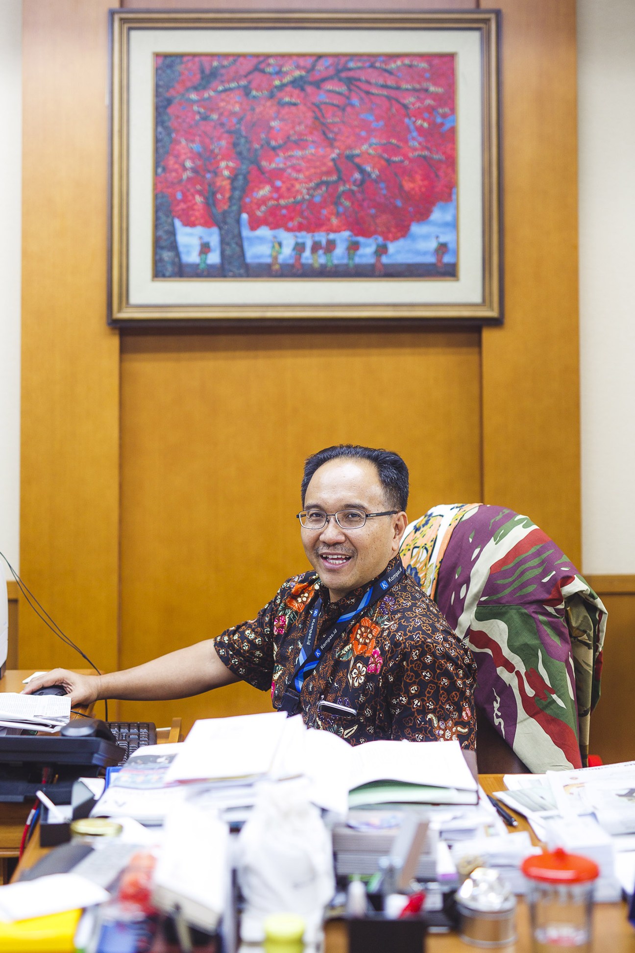 Kompas editor Budiman Tanuredjo seated at his desk in a yellow office with an Indonesian landscape painting above.