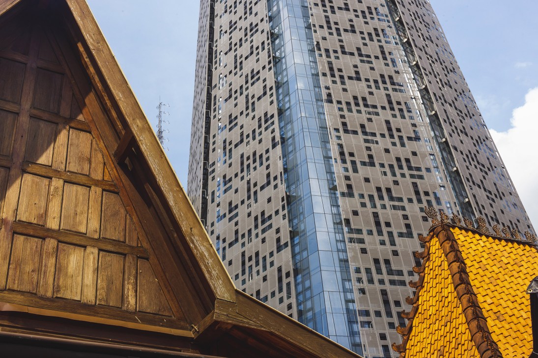 Modern 28-storey grey-green tower building rising above traditional wooden architecture with yellow-tiled roof in Jakarta.