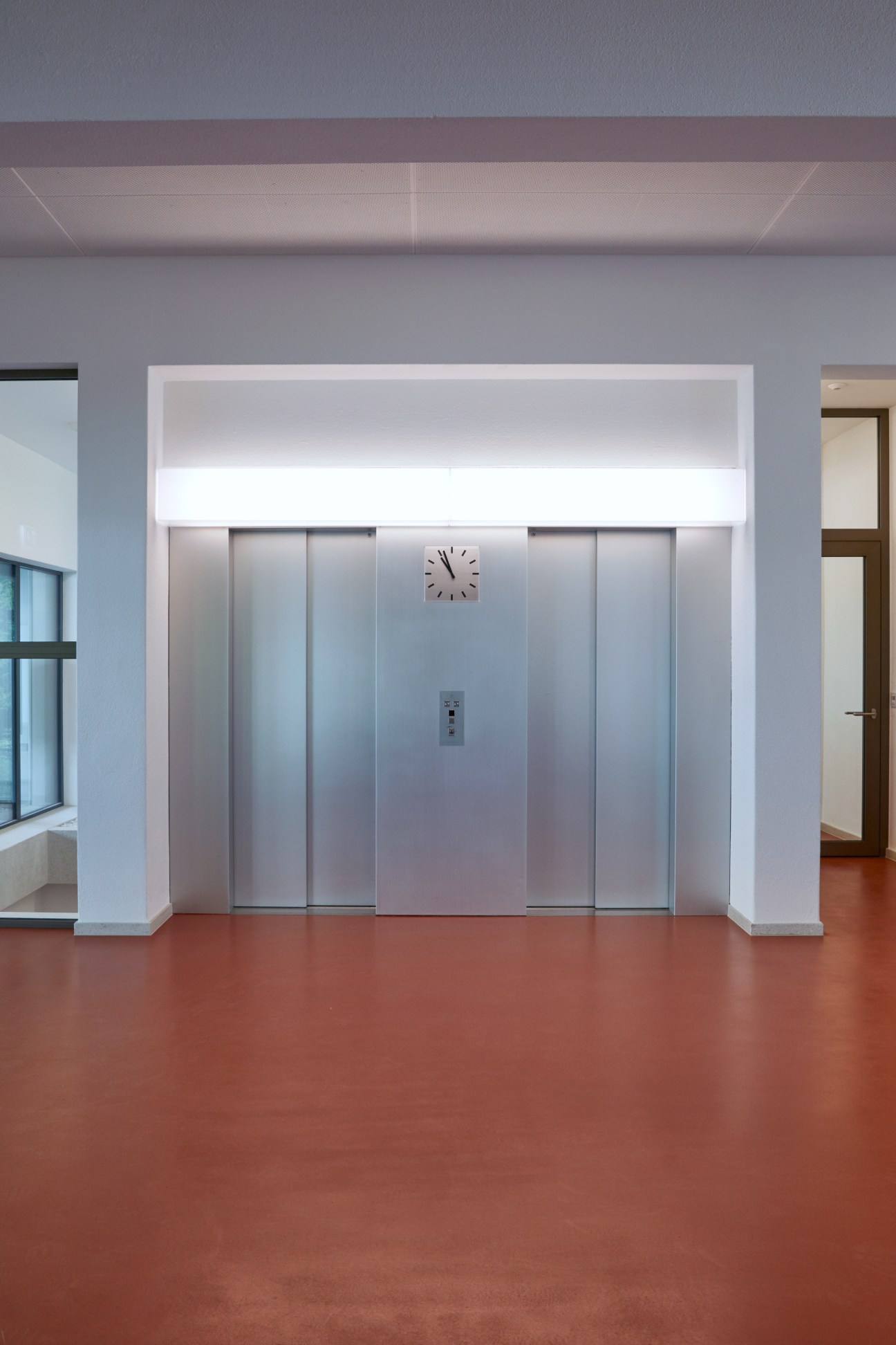 Modern elevator doors with metal panels and a clock mounted above, in a minimalist white corridor with terracotta flooring.
