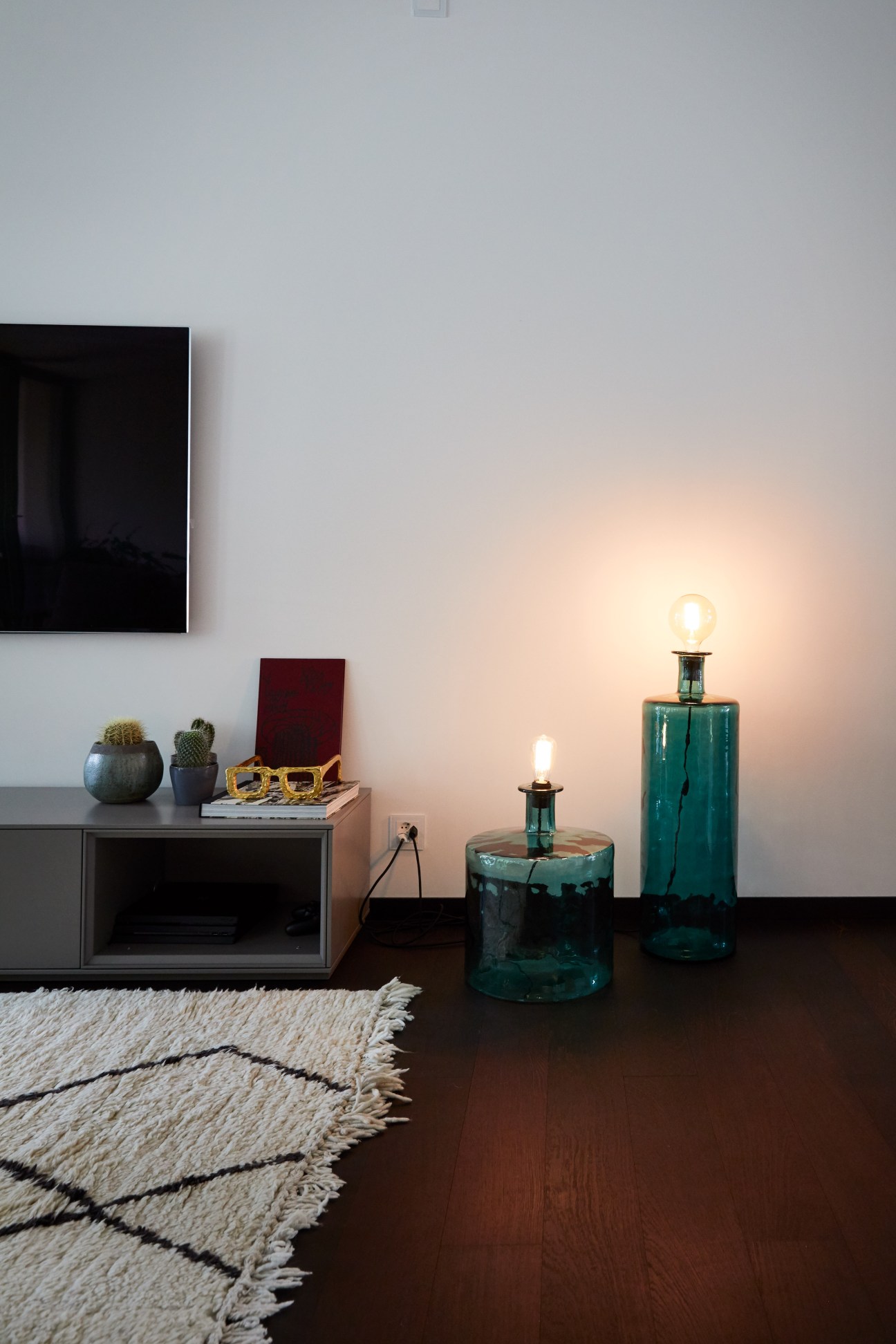 Modern apartment interior with styled shelving, green glass lamps, potted cacti, and geometric rug.