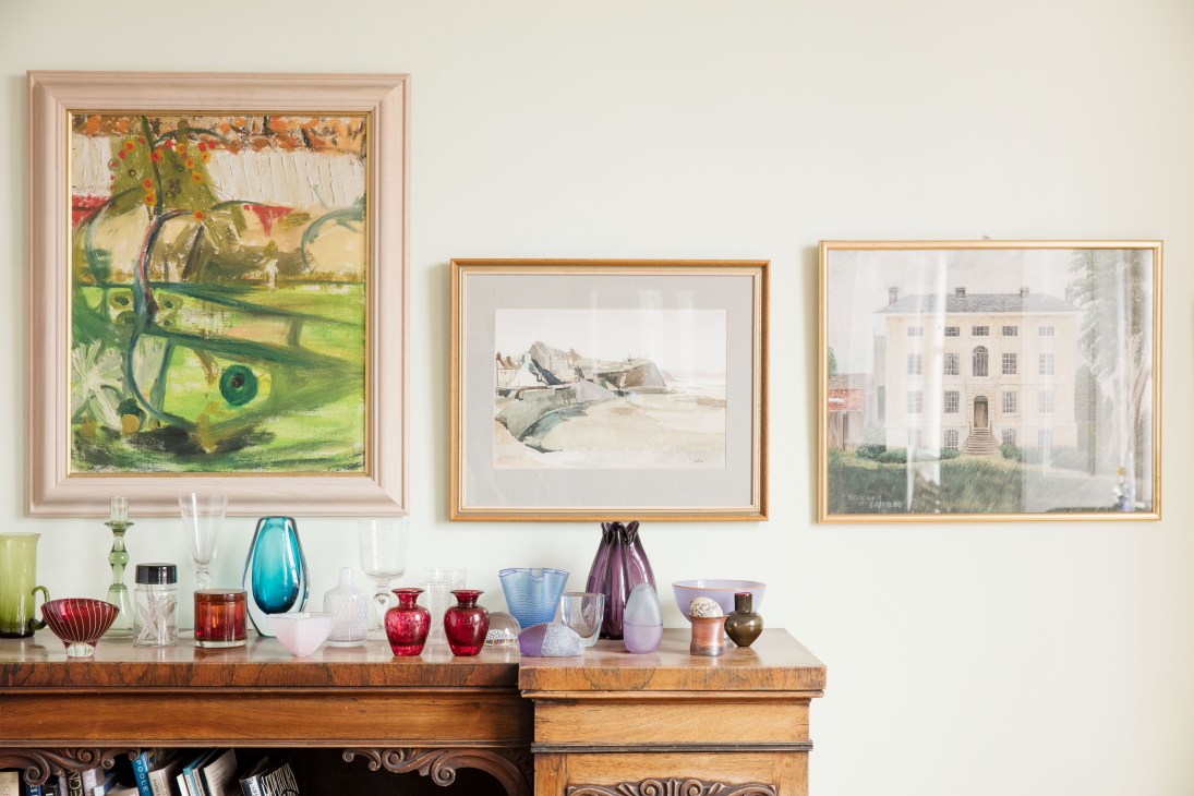 Paintings and colorful mid-century glassware displayed on wooden furniture against a white wall in Ken Stradling's Bristol home.