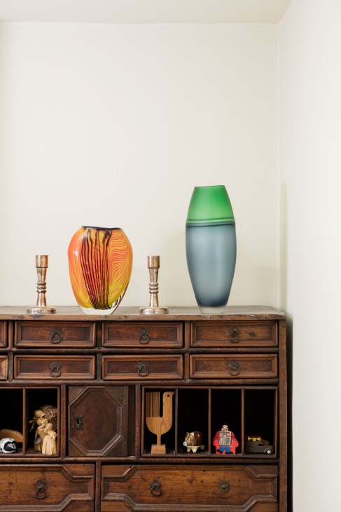 Colorful glass vessels and metallic candlesticks displayed on a wooden cabinet with open compartments below.