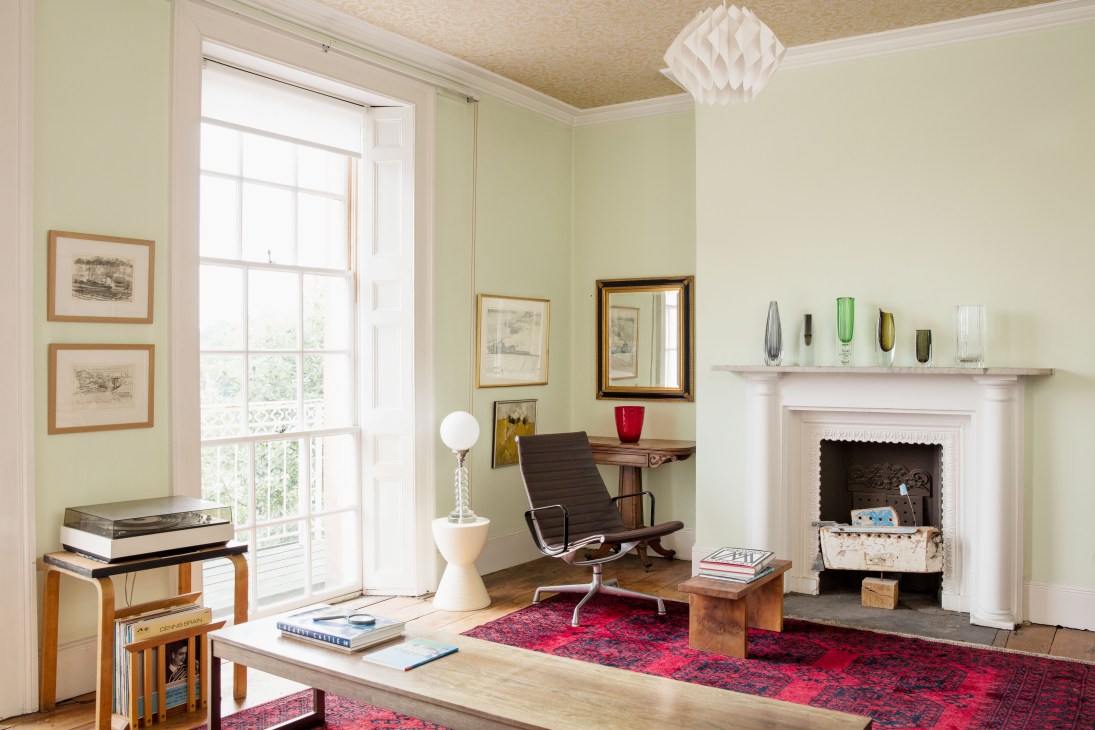 Light-filled modernist sitting room with pale green walls, mid-century furniture, glass collection on shelves, and Afghan carpet.