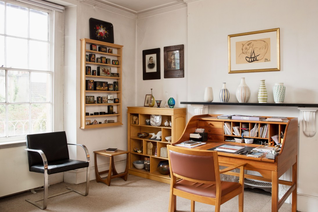 Mid-century modern study featuring wooden desk, shelving units, ceramics, framed artwork, and period furniture by windows.